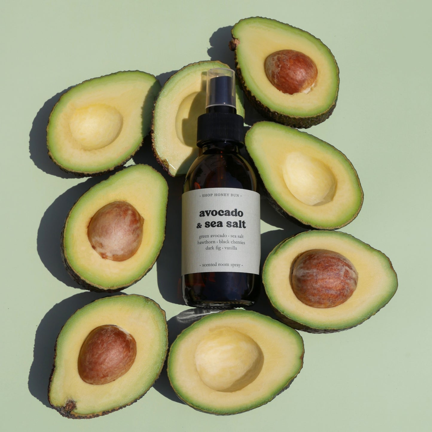 An avocado and sea salt scented room spray surrounded by cut in half avocados on a pale green background