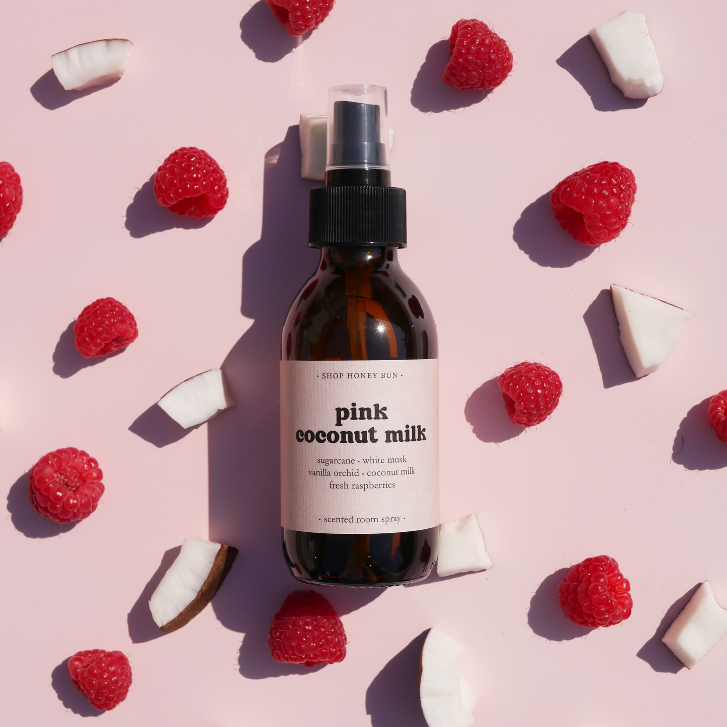 A pink coconut milk scented room spray in an amber glass spray bottle surrounded by coconut and raspberries