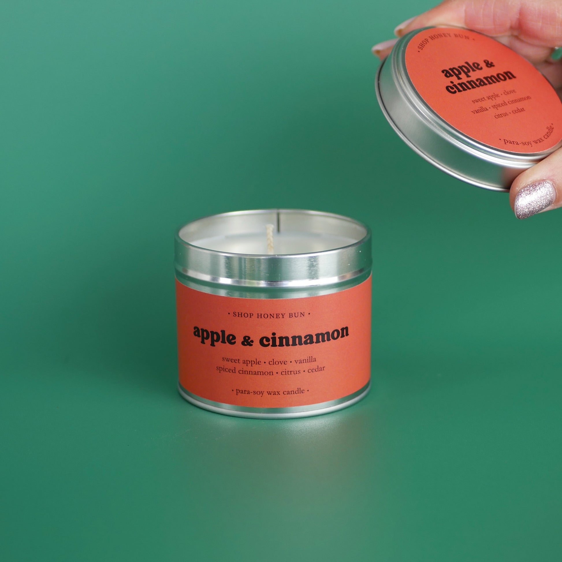 A large apple and cinnamon scented tin candle with a hand holding the lid off to show the candle wick