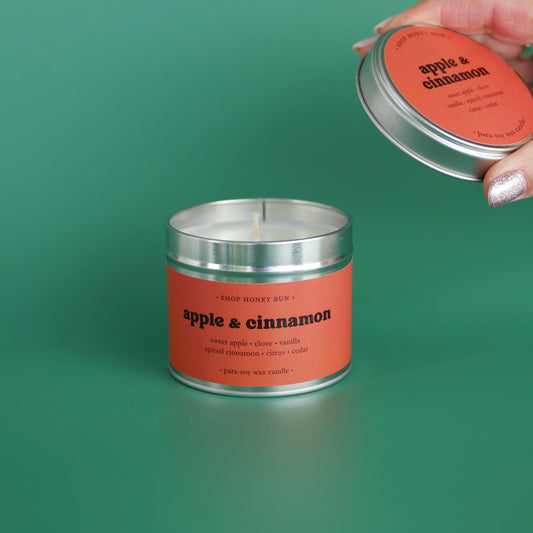 A large apple and cinnamon scented tin candle with a hand holding the lid off to show the candle wick