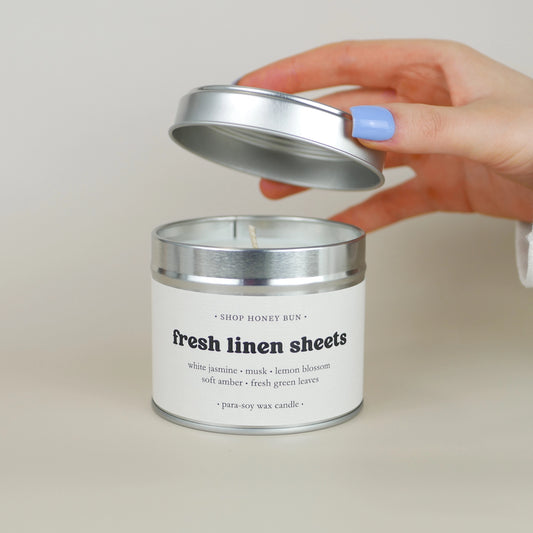 A 30 hour burn time fresh linen sheets scented in candle with a hand lifting off the lid to show the candle wick
