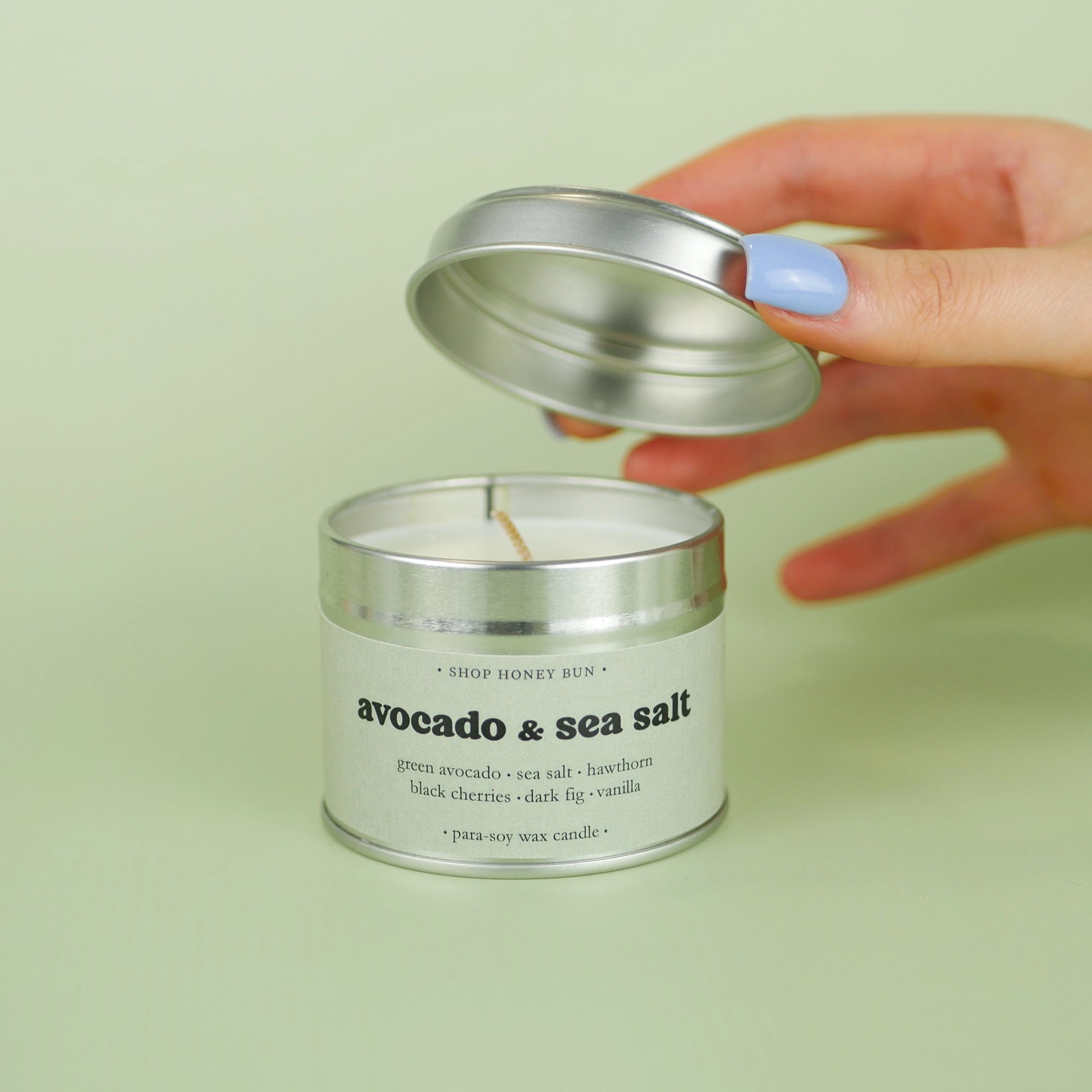 A 20 hour burn time avocado and sea salt scented in candle with a hand lifting off the lid to show the candle wick