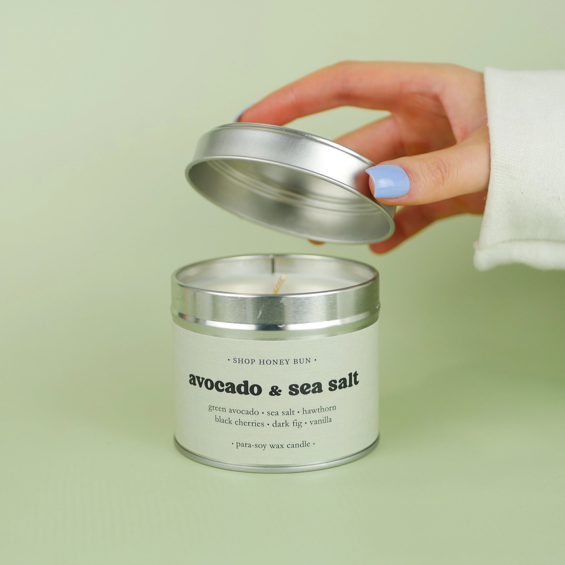 A 30 hour burn time avocado and sea salt scented in candle with a hand lifting off the lid to show the candle wick