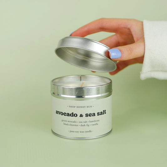 A 30 hour burn time avocado and sea salt scented in candle with a hand lifting off the lid to show the candle wick