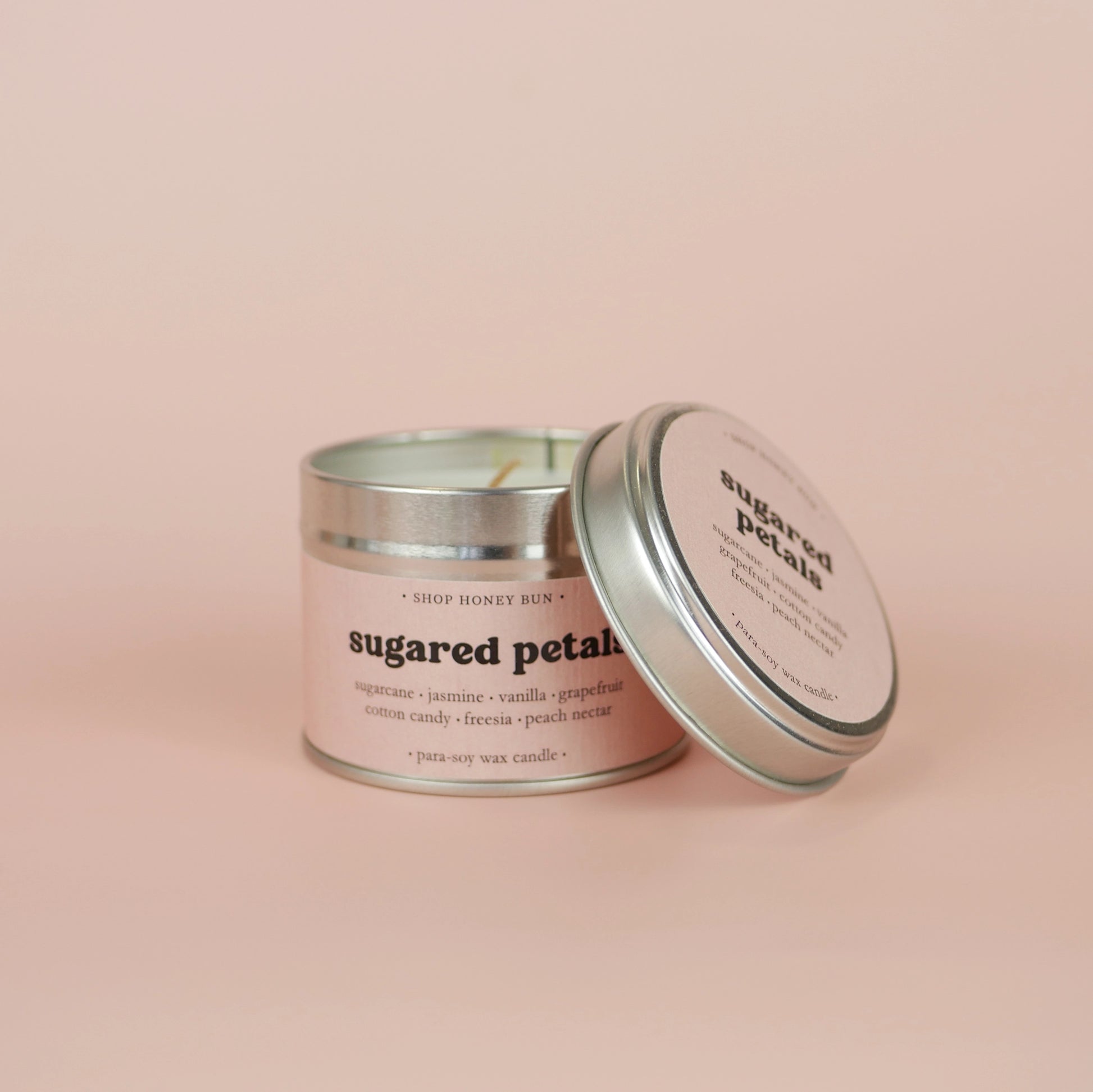 A 20 hour burn time sugared petals scented tin candle with the lid off, resting on the side of the candle
