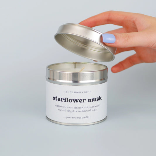 A 30 hour burn time starflower musk scented in candle with a hand lifting off the lid to show the candle wick
