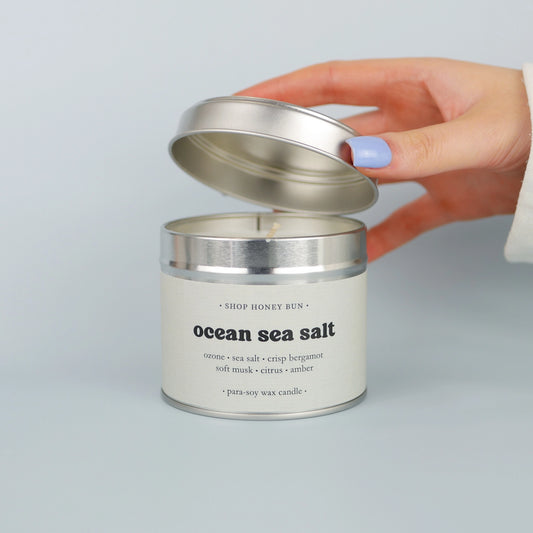 A 30 hour burn time ocean sea salt scented in candle with a hand lifting off the lid to show the candle wick